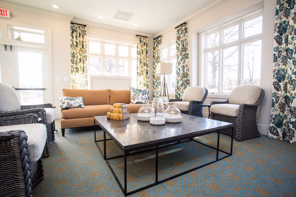 A bright and cozy living room with large windows letting in natural light. The room features a brown sofa with patterned cushions, two wicker armchairs with light cushions, a large square coffee table with decorative items, and floral curtains. The carpet is blue with a subtle pattern, and there is a floor lamp in the corner near the windows.