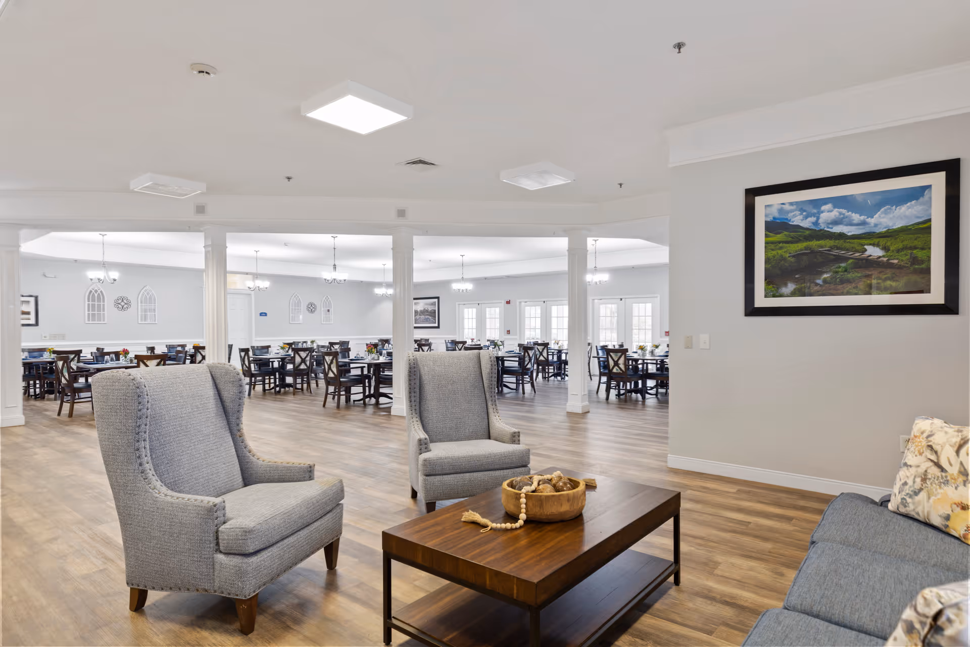 Open common area with two gray armchairs and a coffee table in the foreground and a large dining room with tables and chairs in the background.