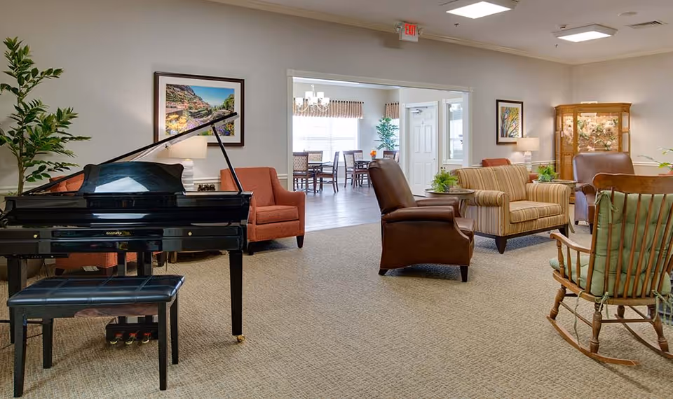A spacious senior living common area featuring a black grand piano with a matching bench, various comfortable chairs including a striped loveseat, leather armchair, and wooden rocking chair. The room is decorated with framed artwork, lamps, plants, and a wooden display cabinet. In the background, a dining area with tables and chairs is visible through a wide doorway.