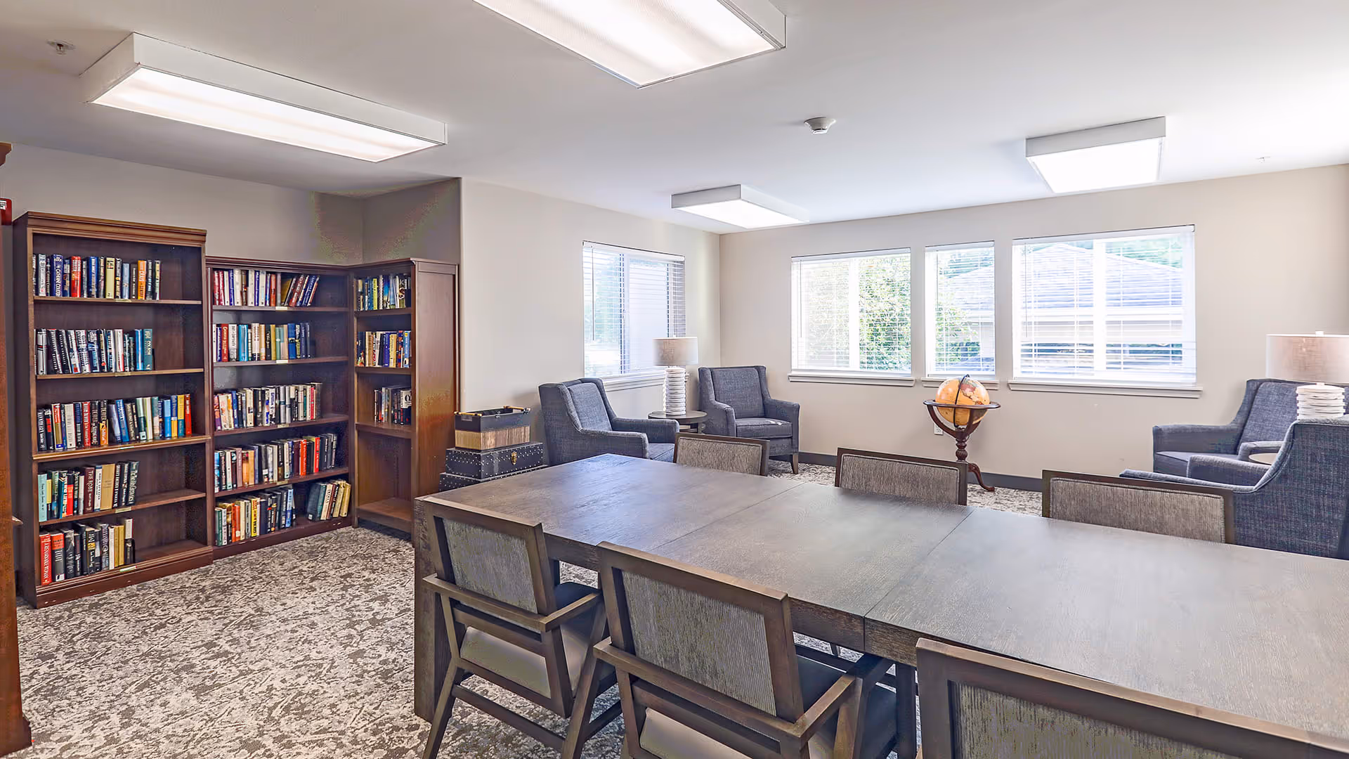 A bright and spacious common room with a large wooden table surrounded by chairs in the foreground. In the background, there are three armchairs arranged near windows with white blinds, a globe on a stand, and a corner bookshelf filled with books. The room has carpeted flooring and ceiling lights.