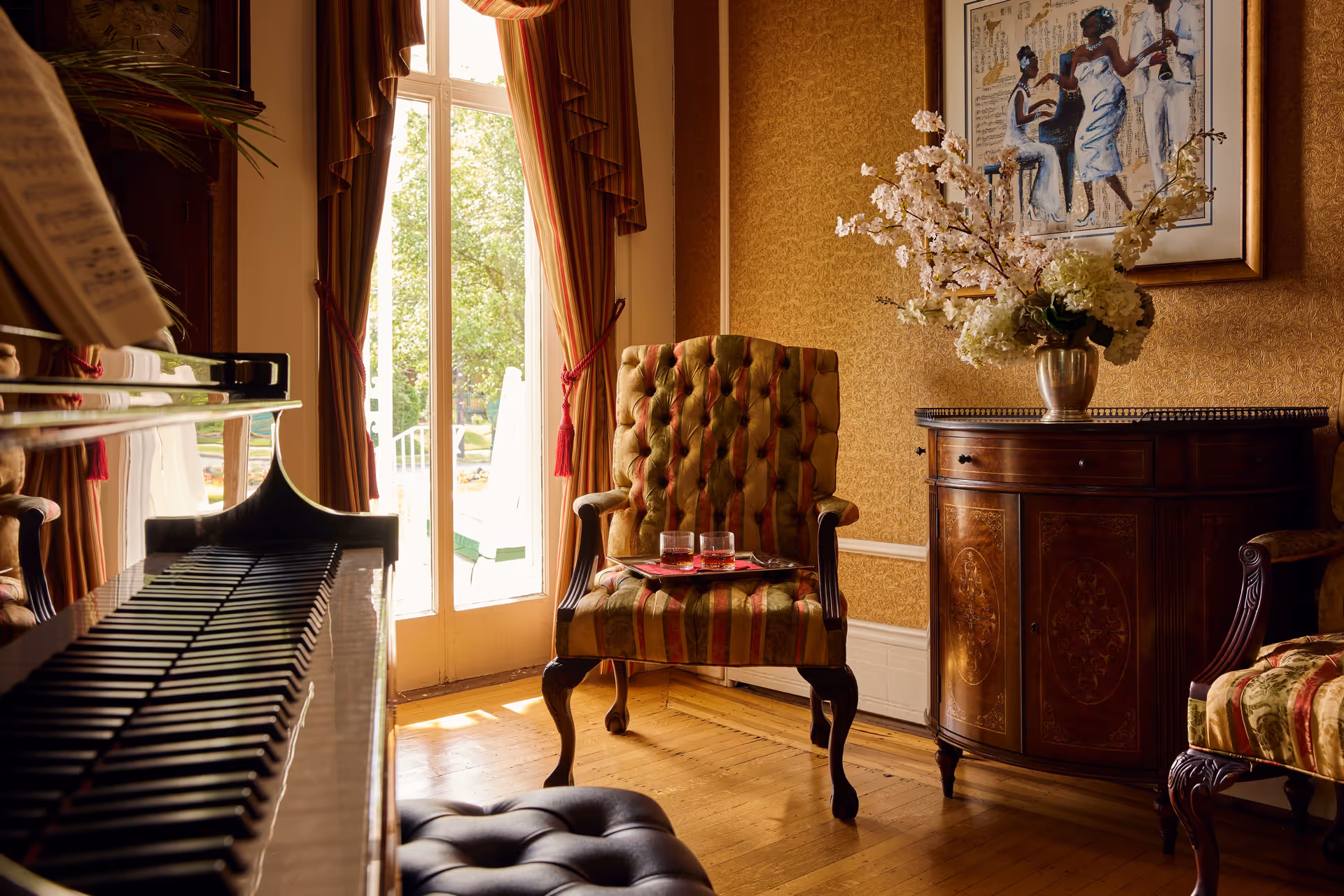 A cozy, elegant living room area with a polished piano on the left, a striped upholstered armchair with two glasses on a tray, a wooden cabinet with a vase of flowers, and a framed painting of musicians on the wall. Sunlight streams through glass doors leading to an outdoor garden.