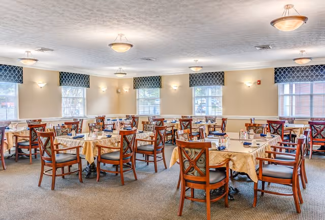 A bright dining room with multiple tables covered in beige tablecloths, each surrounded by wooden chairs with cushioned seats. The room has several windows with blue patterned valances, beige walls, and ceiling lights providing warm illumination.