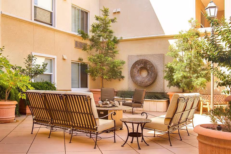 Outdoor patio area with cushioned metal chairs and sofas arranged around a small round table and a fire pit. The space is surrounded by potted plants and trees, with a beige building wall featuring windows and a circular wall fountain in the background. A lamp post is visible on the right side.
