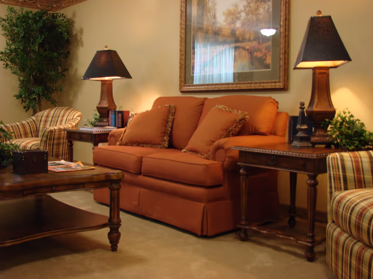 A cozy living room with an orange sofa adorned with matching pillows, two wooden side tables each holding a lamp with a dark shade, a wooden coffee table with magazines and a small chest, two striped armchairs, a large framed landscape painting on the wall, and a tall green plant in the corner.