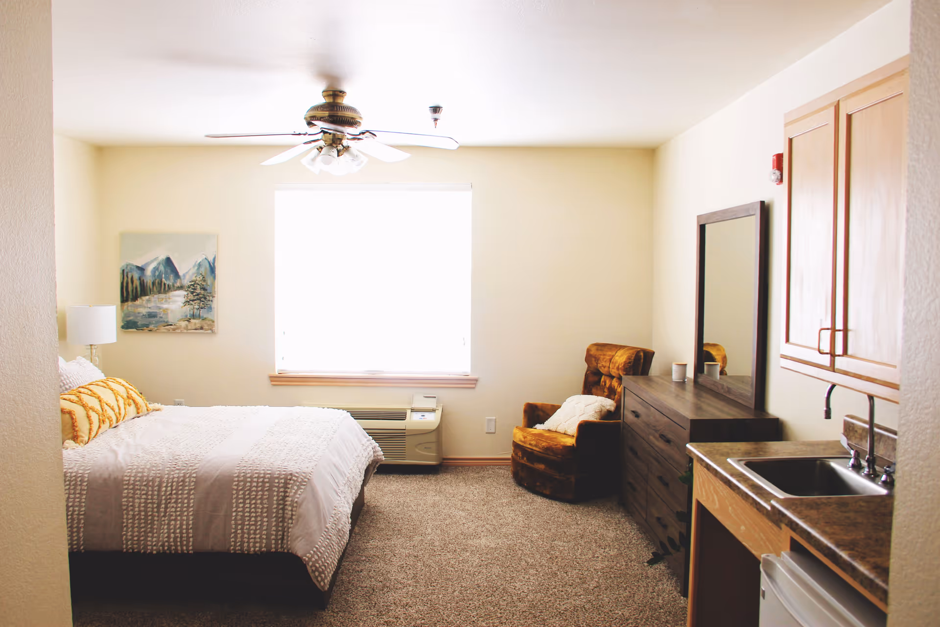 A cozy bedroom with a large bed covered in white and beige bedding, a yellow patterned pillow, a bedside lamp, a painting of mountains on the wall, a ceiling fan, a window with bright light coming through, a brown armchair with a white pillow, a dark wooden dresser with a mirror, and a small kitchenette area with a sink and cabinets.