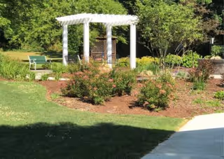 A garden area with a white pergola structure surrounded by green bushes and plants. There is a green bench on the left side and a concrete pathway on the right. Trees and other greenery are visible in the background.