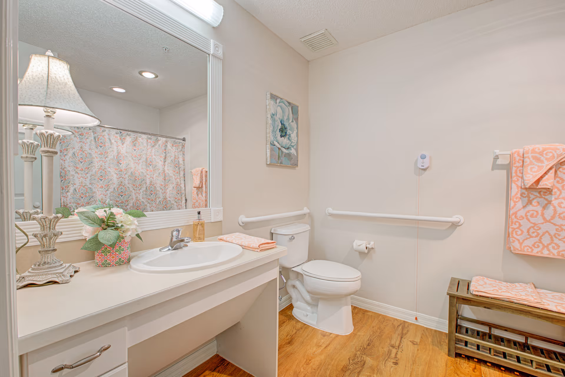 A clean and well-lit bathroom featuring a white sink with a countertop, a large mirror, a decorative lamp, and a small flower arrangement. There is a toilet with grab bars on the wall for accessibility, a towel rack with folded orange patterned towels, and a wooden bench. The floor has a wood-like finish and the walls are painted light beige.