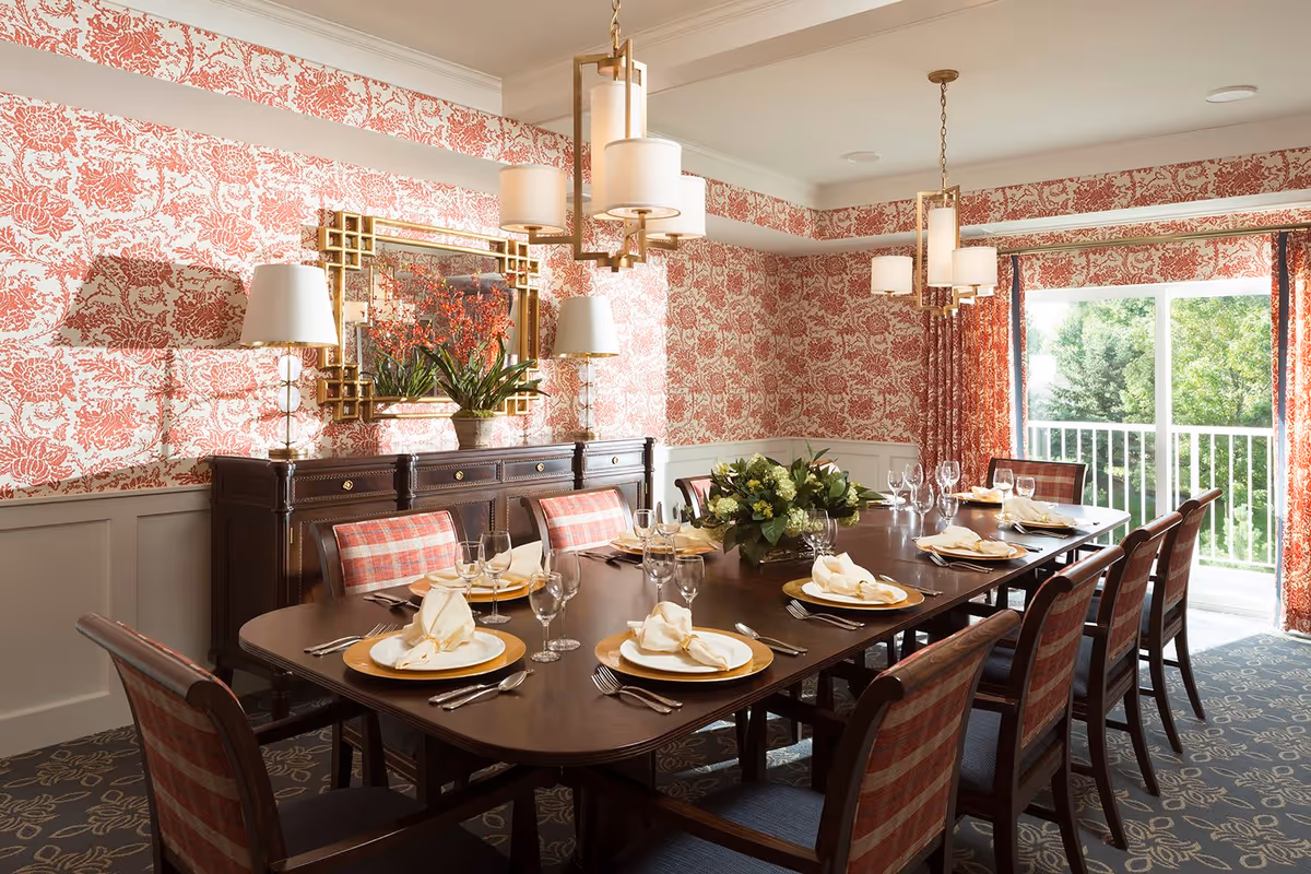 Elegantly set dining room with a long table, place settings, patterned red wallpaper, and a balcony view.