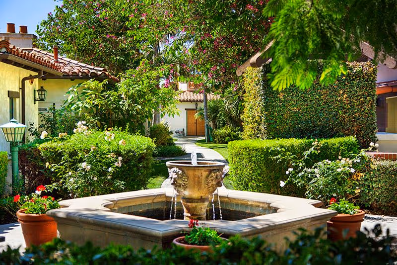 Sunny landscaped courtyard with a central stone fountain, potted flowers, trimmed hedges, and Spanish-style buildings in the background.