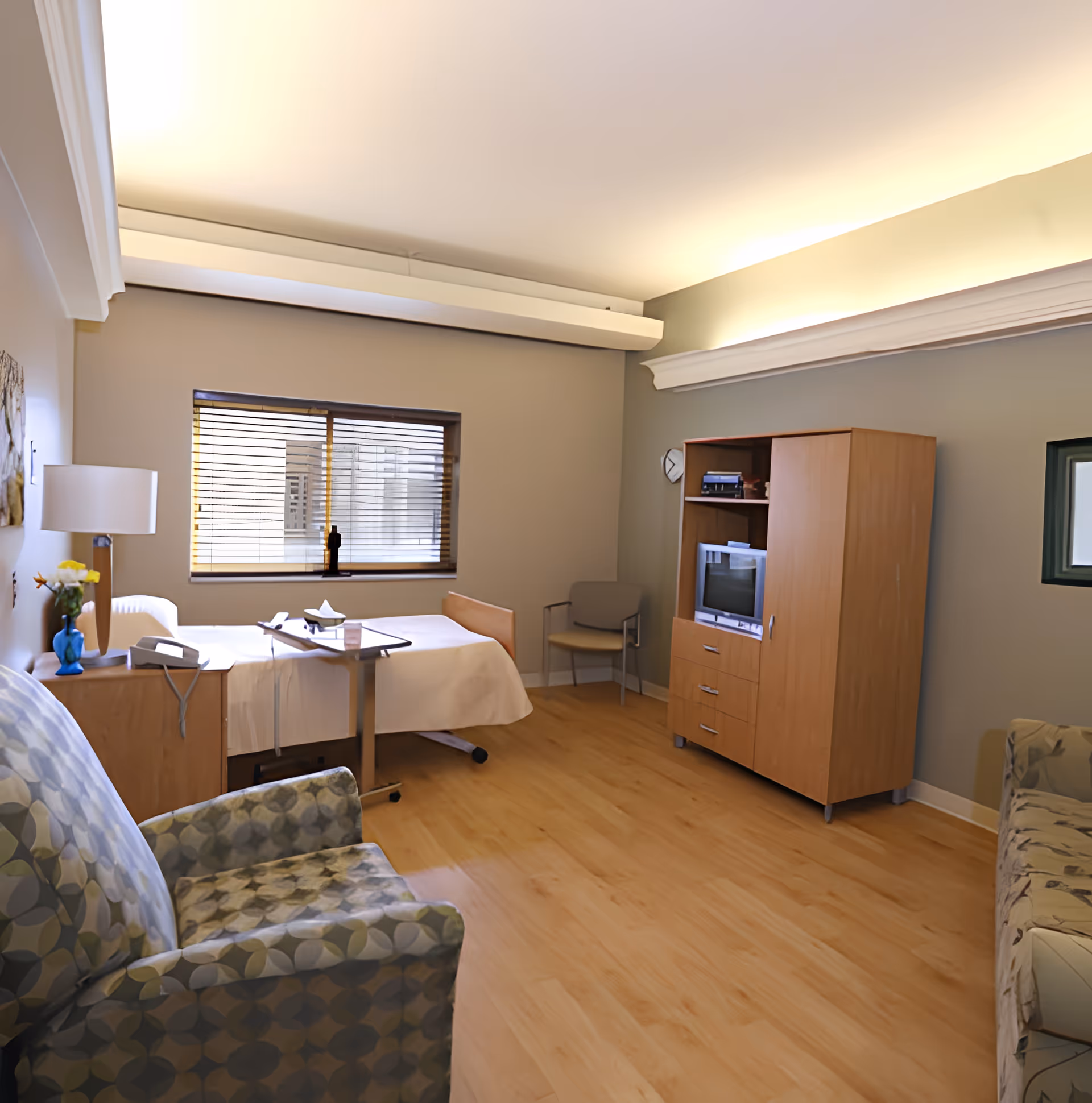 A well-lit room with a single hospital-style bed, a bedside table with a lamp and flowers, a patterned armchair, a wooden cabinet with a TV, and a window with blinds. The room has light-colored walls and wooden flooring.