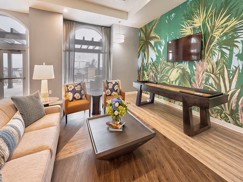 A bright and inviting living room area with a beige sofa adorned with patterned pillows, two orange armchairs with floral cushions, a dark wooden coffee table with a vase of flowers and books, and a shuffleboard table against a wall featuring a large tropical mural. Large windows with sheer curtains allow natural light to fill the room.