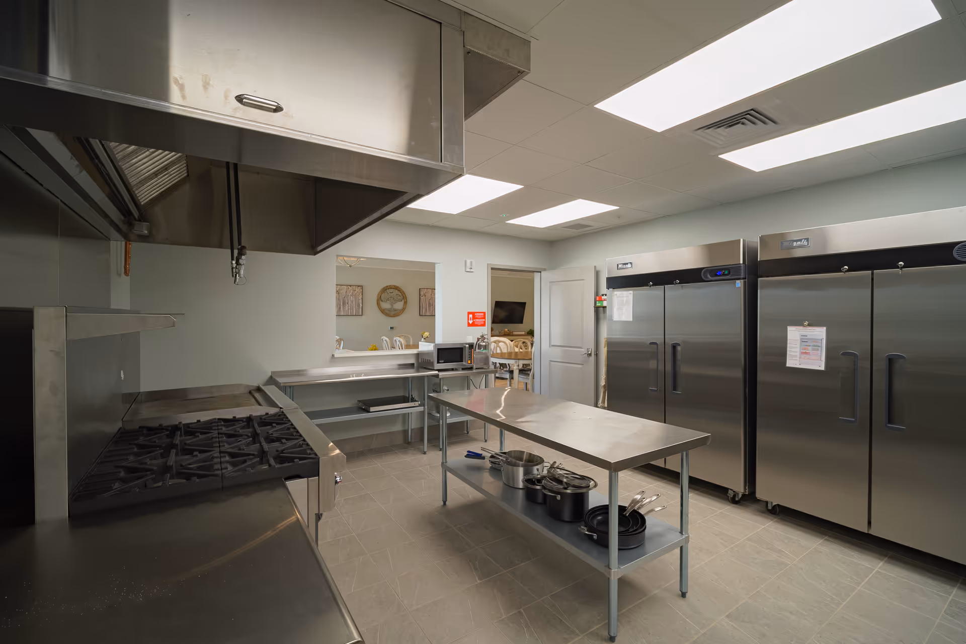 A clean commercial kitchen with stainless steel appliances including a stove, two large refrigerators, a microwave, and a central stainless steel work table with pots and pans underneath. The kitchen has tiled flooring and bright overhead lighting. Through an open doorway, a dining area with chairs and wall decorations is visible.