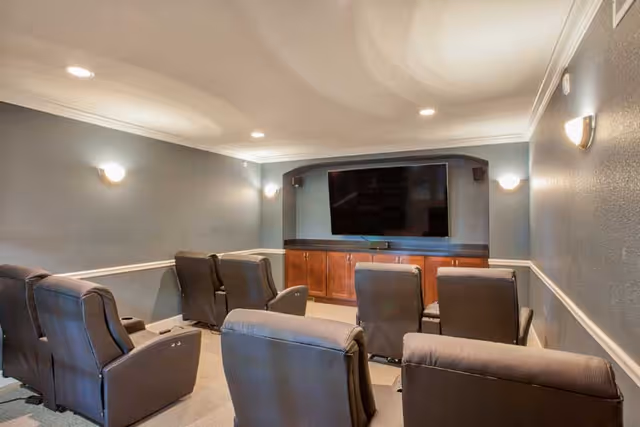 A small theater room with six black recliner chairs arranged in two rows facing a large flat-screen TV mounted on a wall with wooden cabinetry below. The room has gray walls with white trim and recessed ceiling lights.