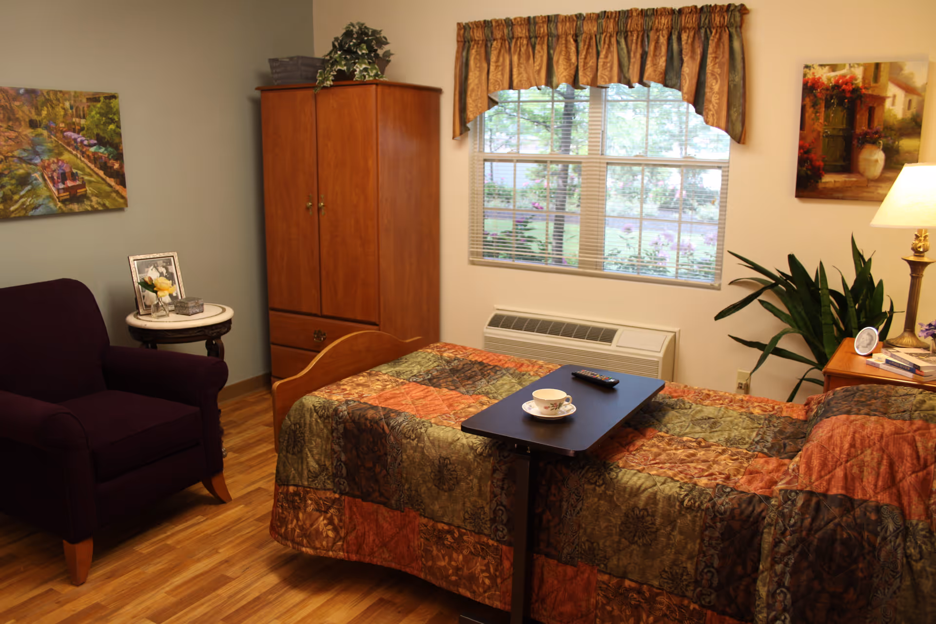 A cozy bedroom with a single bed covered in a multicolored quilt. A small black overbed table holds a cup and saucer and a remote control. There is a wooden armoire against the wall, a purple armchair next to a small round table with a framed photo and decorative items. A window with blinds and a valance lets in natural light, and there are paintings on the walls and a potted plant near a wooden nightstand with a lamp and books.