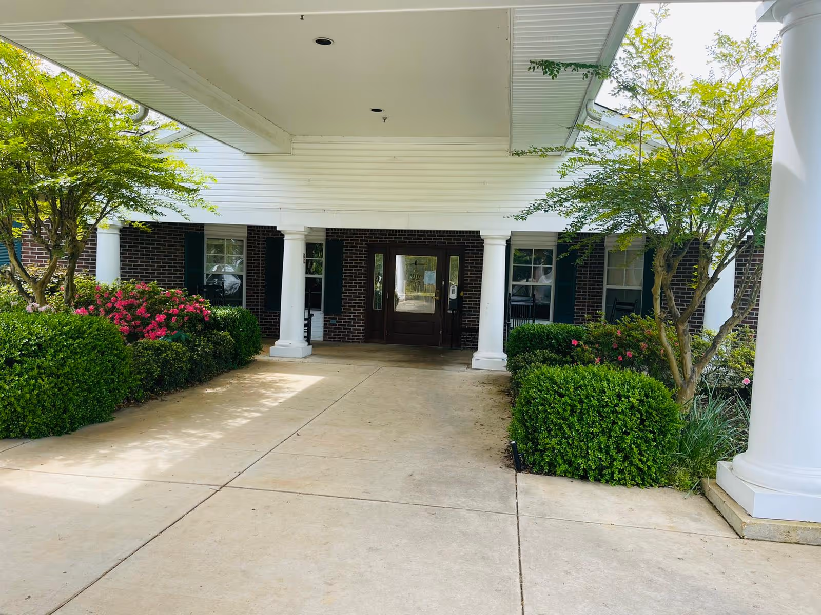Covered entrance to a building with white columns and a concrete driveway. There are green bushes and flowering plants on either side of the entrance, and the building has dark brick walls with windows and dark shutters.