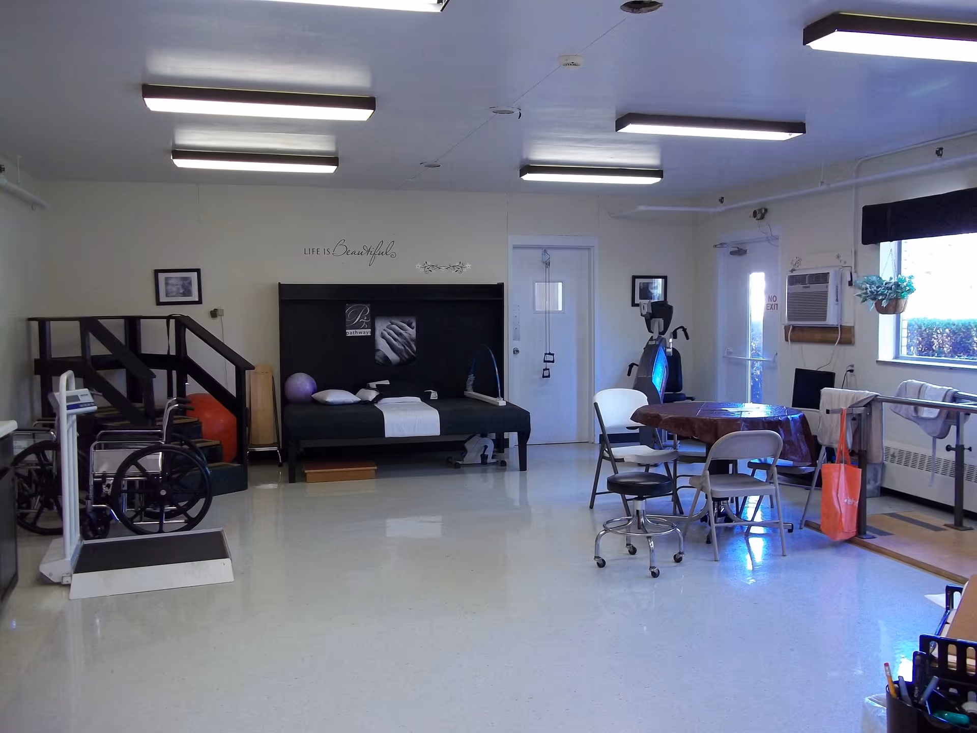 A spacious therapy or rehabilitation room with exercise equipment including a wheelchair, parallel bars, and a therapy bed. There is a round table with chairs in the center, a window with a hanging plant, and motivational wall decor that reads 'Life is Beautiful'. The room is well-lit with fluorescent ceiling lights.