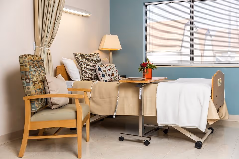A hospital or rehabilitation room with a bed covered in beige and white linens, several patterned pillows, a wooden chair with a cushion, a small table on wheels with a flower arrangement, a lamp, and a window with blinds.