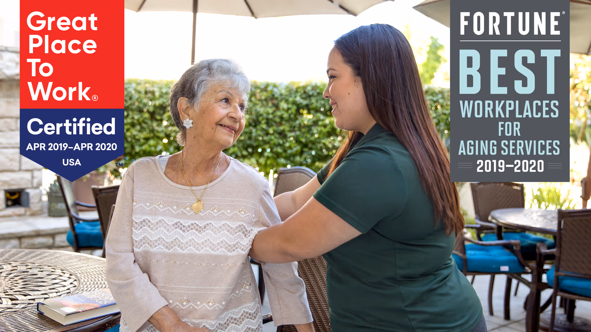 An elderly woman sitting outdoors at a table with a younger caregiver smiling and holding her arm. There are patio umbrellas and chairs in the background, with greenery behind them.