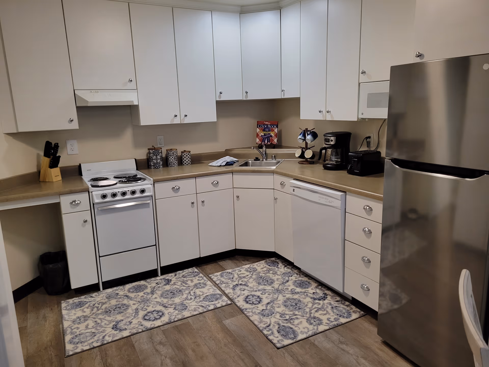 A clean and modern kitchen with white cabinets and beige countertops. The kitchen features a white electric stove, a stainless steel refrigerator, a dishwasher, a microwave, a coffee maker, a toaster, and a knife block. There are two patterned rugs on the wooden floor and decorative canisters on the counter near the stove.