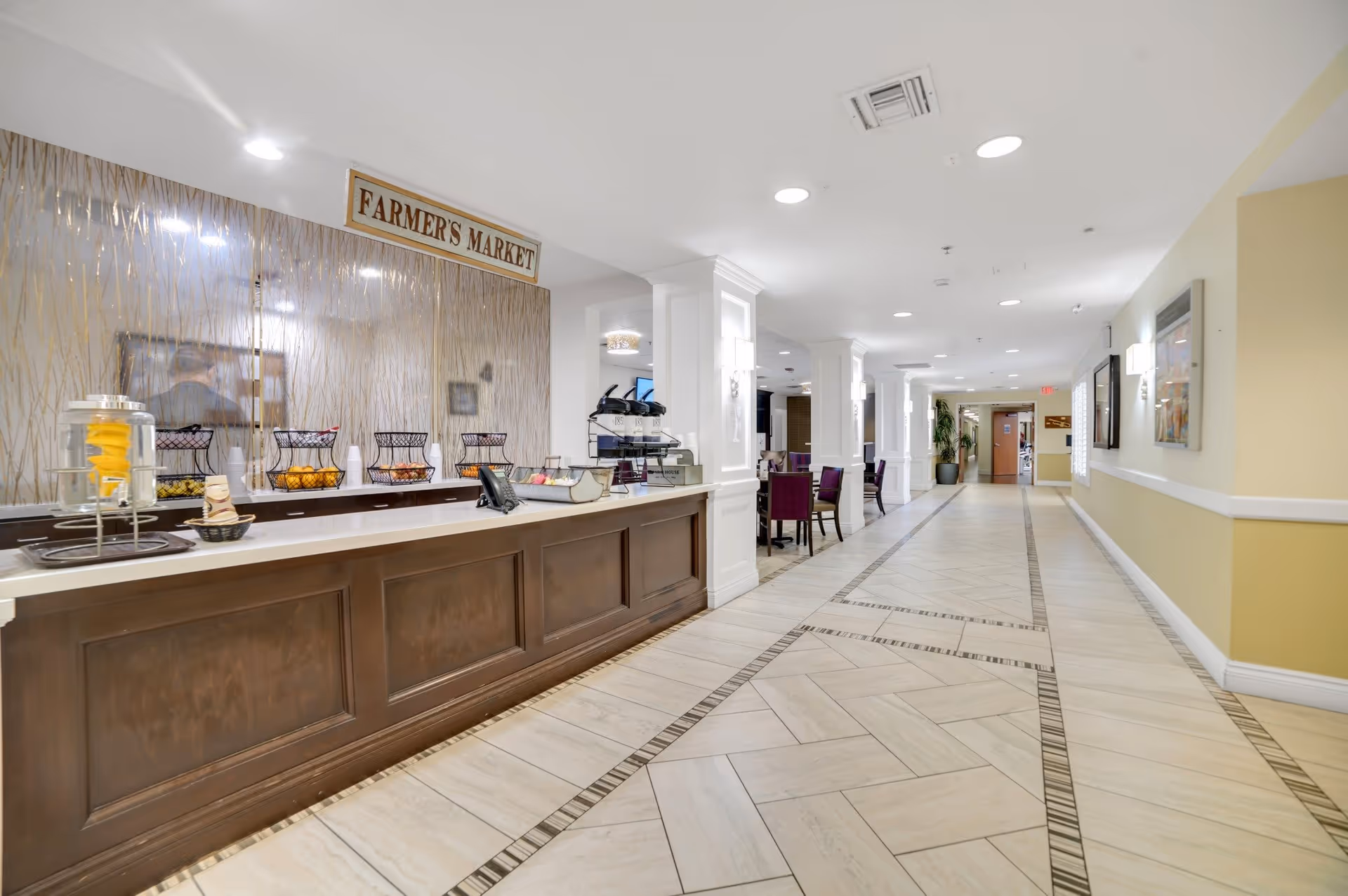 Interior hallway of a senior living facility with a long wooden counter labeled 'Farmer's Market' on the left side, featuring baskets of snacks and a water dispenser. The hallway has tiled floors with decorative patterns, white walls with yellow accents, and recessed lighting. There are tables and chairs along the hallway and framed artwork on the walls.