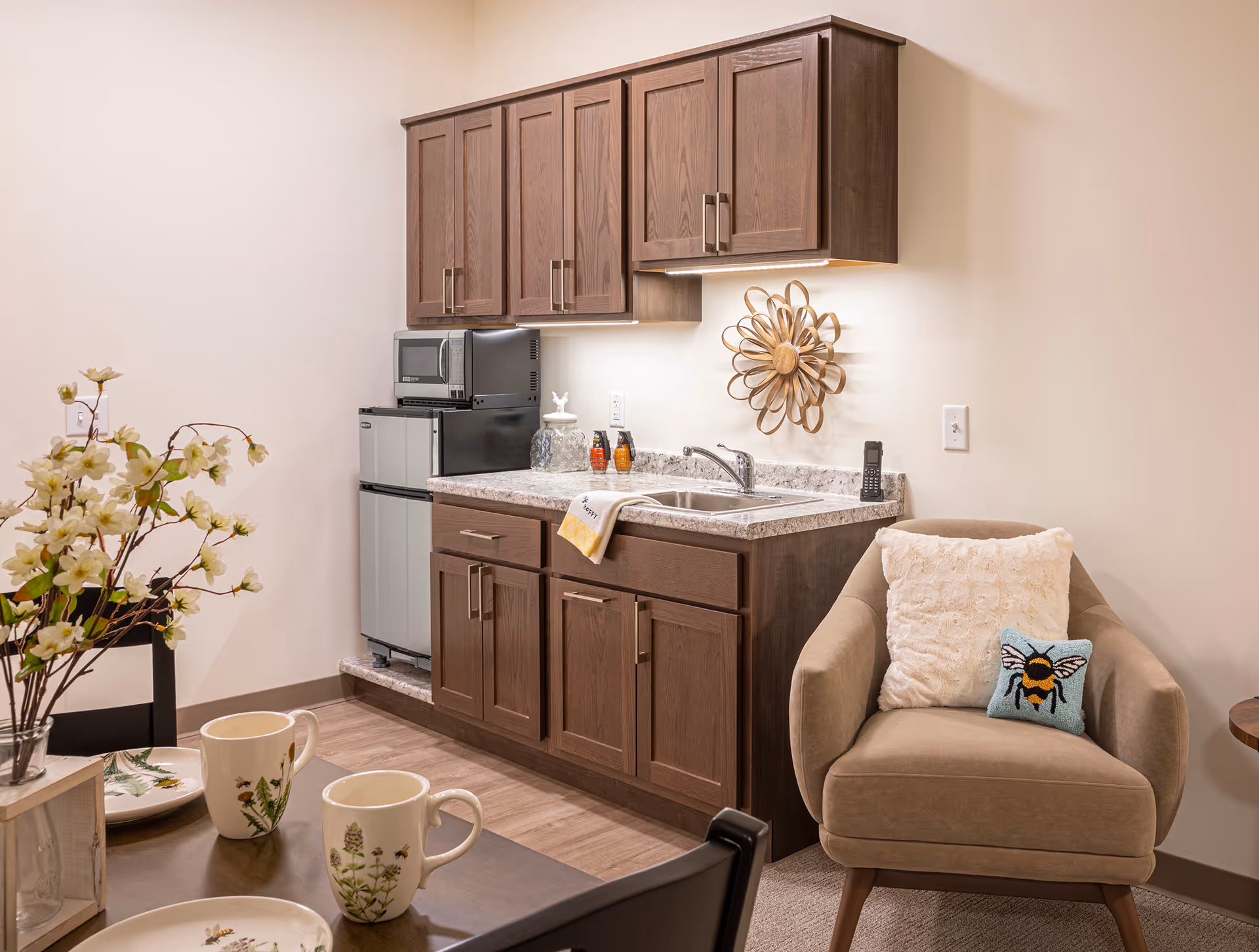 Small kitchenette with dark wood cabinets, a sink and mini fridge next to a cozy armchair and a set dining table.