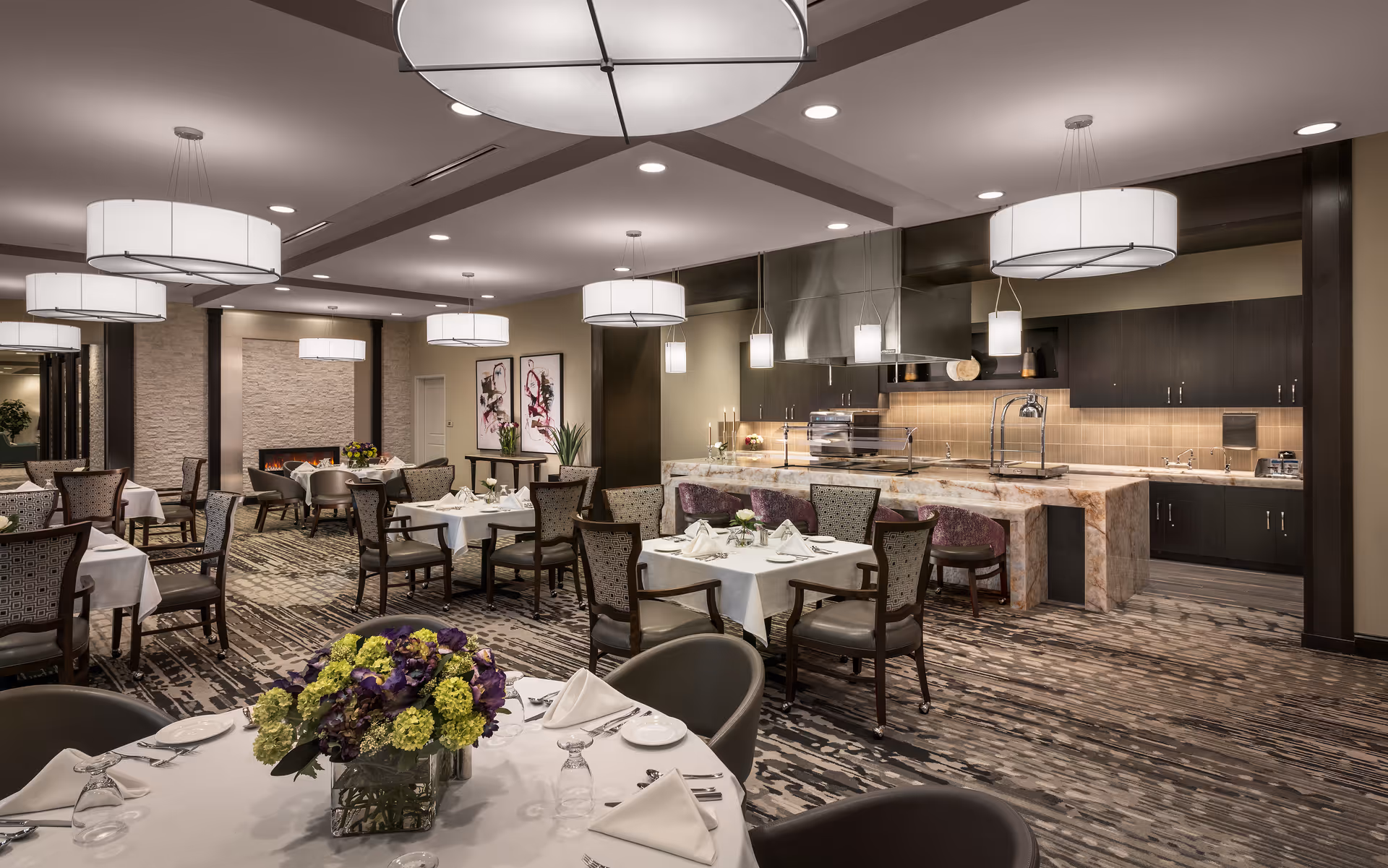 A spacious, elegantly decorated dining room with multiple tables covered in white tablecloths, set with plates, glasses, and napkins. The room features modern pendant lighting, a fireplace, floral centerpieces on the tables, and a large kitchen area with a marble countertop and dark cabinetry in the background.