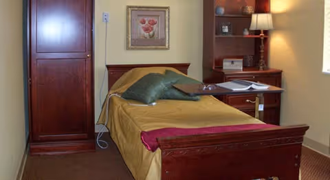 A single bed with a wooden headboard and footboard in a room. The bed is made with a yellow bedspread, a green pillow, and a red blanket at the foot. Next to the bed is a wooden nightstand with a lamp, a book, and some decorative items. A wooden wardrobe and a framed flower picture are on the wall behind the bed. The room has beige walls and carpeted flooring.