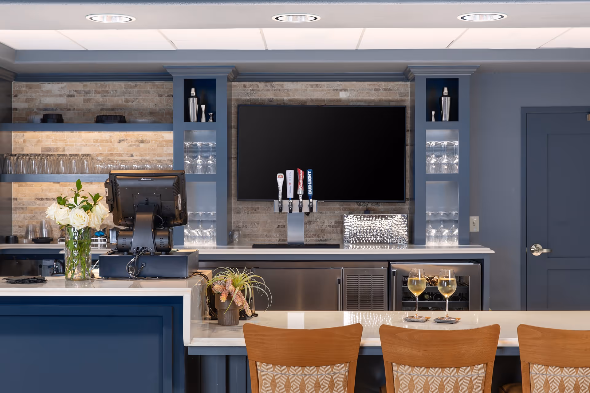 Interior view of a modern bar area with a blue and white color scheme. The bar counter has three wooden chairs with patterned cushions, two glasses of white wine on coasters, and a small plant. Behind the bar is a large flat-screen TV mounted on a stone-tiled wall, three beer taps, shelves with glassware, and a cash register on the counter.