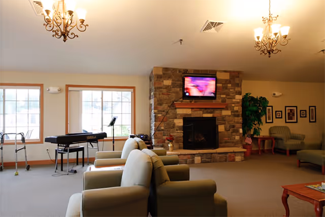 A cozy assisted living common area with several cushioned armchairs arranged facing a stone fireplace with a mounted flat-screen TV above it. There is a keyboard and music stand near large windows letting in natural light. The room is warmly lit by chandeliers and decorated with framed pictures and a potted plant.