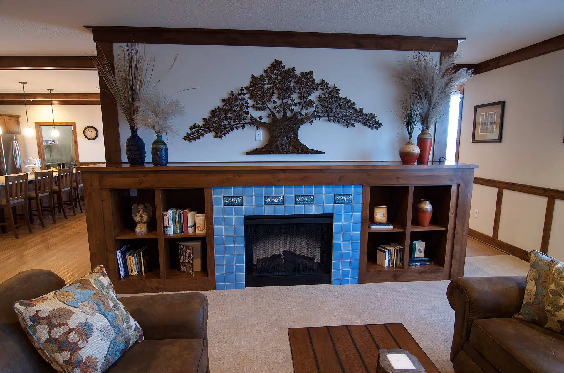 A cozy living room area featuring a fireplace with blue tile surround and wooden built-in shelves on either side holding books and decorative items. Above the fireplace is a metal wall art piece shaped like a tree. Two armchairs with floral patterned pillows face a wooden coffee table. The room has wooden trim and a view into a dining area with a long table and chairs.