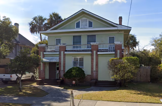 Exterior front view of a two-story residential building with a green facade and red brick accents. The house has a small porch with an awning over the entrance, several windows, and a balcony on the second floor. There are trees and bushes in the yard, and a white pickup truck is parked on the left side.