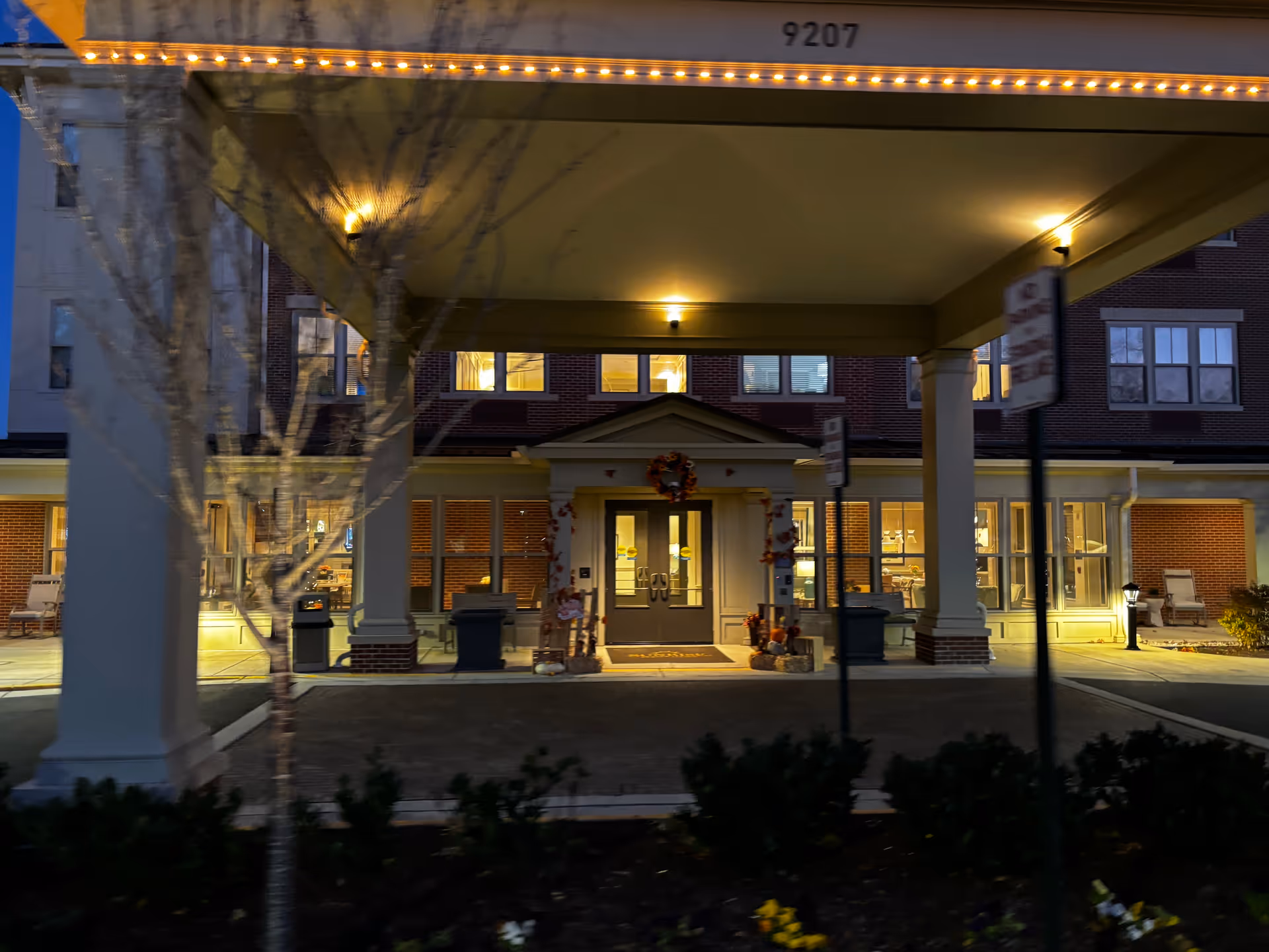 Lit covered entrance and porte-cochere of a senior living building at dusk with wreath-decorated doors and illuminated windows.