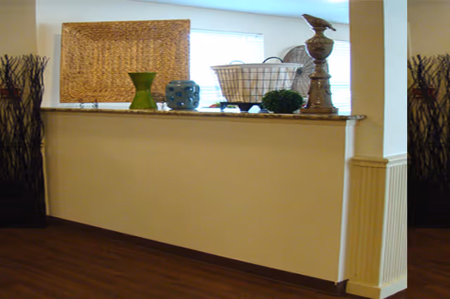 Interior view of a room with a half wall decorated with various items including a green vase, a blue ceramic piece, a wire basket, a small green plant, and a decorative bird sculpture. The floor is wooden, and there are tall decorative sticks in black containers on either side of the wall.
