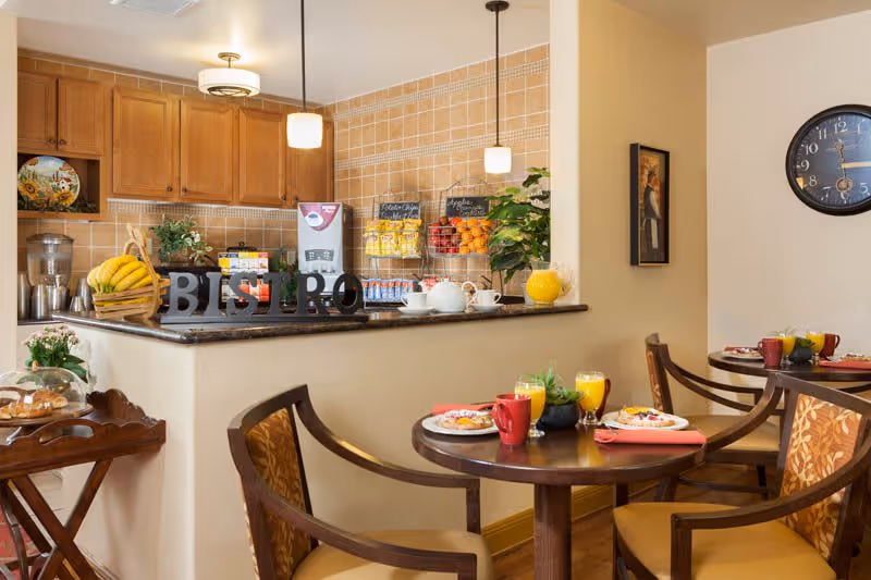 A cozy bistro area with a small round dining table set with plates of food, glasses of orange juice, and red mugs. The background features a kitchen counter with a basket of bananas, a coffee machine, snacks on display racks, and decorative plants. Wooden chairs with patterned cushions surround the tables, and a wall clock and framed artwork are visible on the wall.