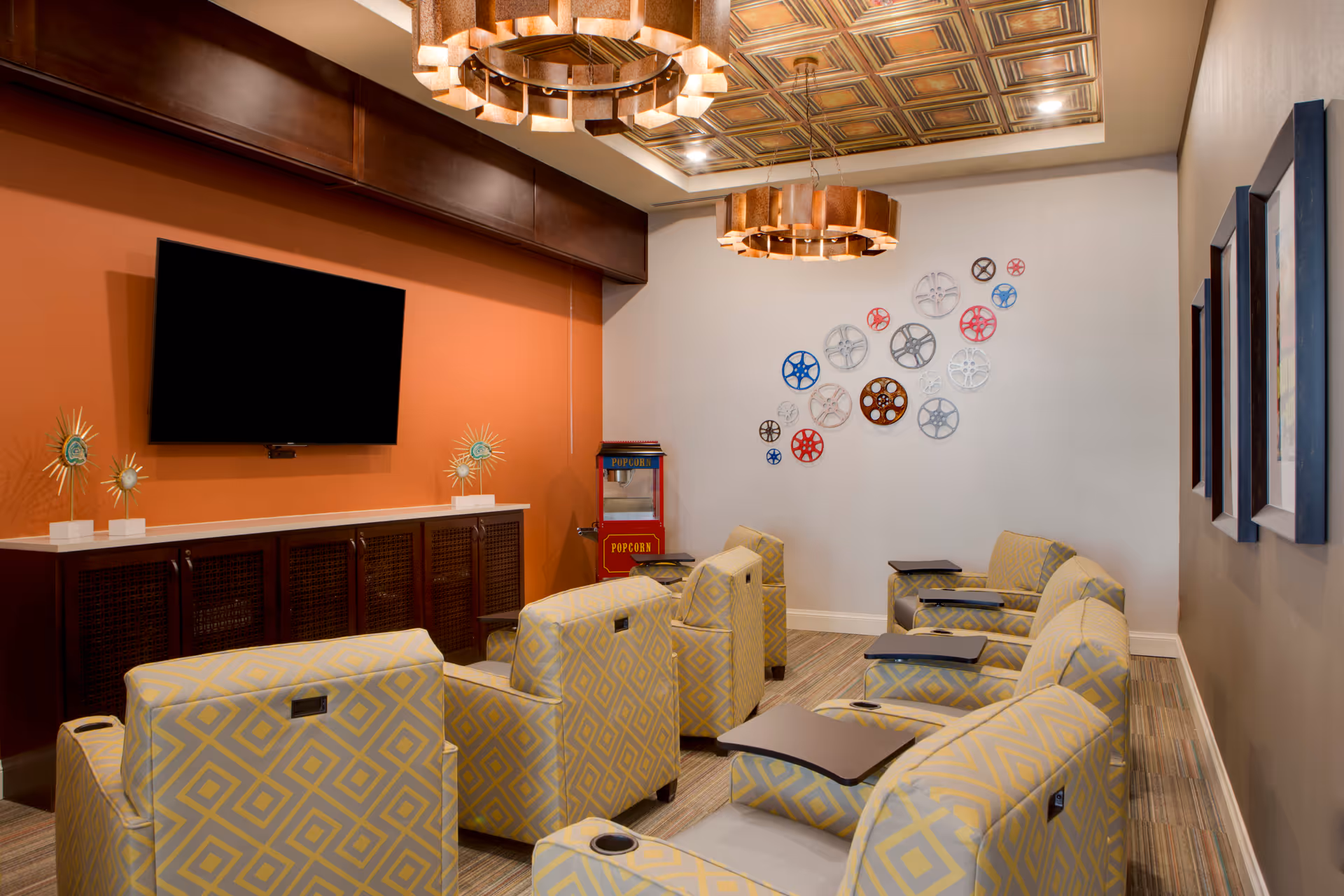 A cozy media room with six patterned reclining chairs arranged in two rows facing a wall-mounted flat screen TV. The room features warm orange and beige walls, decorative metal wall art resembling gears, a popcorn machine in the corner, and two circular pendant lights hanging from a decorative ceiling.