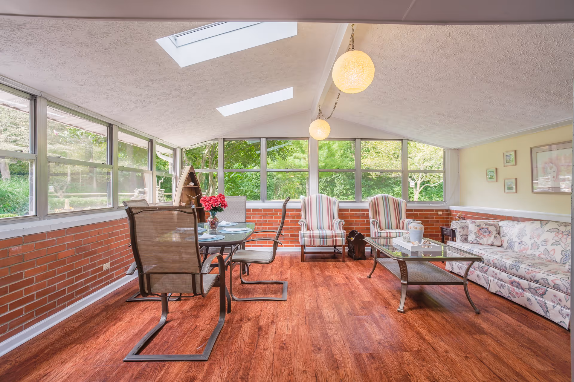 A bright sunroom with large windows and skylights letting in natural light. The room features a glass dining table with four mesh chairs, two striped armchairs, a floral-patterned sofa, and a glass coffee table. The walls have a half-brick design with light yellow paint above, and there are two round pendant lights hanging from the ceiling. Outside greenery is visible through the windows.