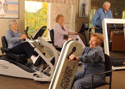 Three elderly women exercising on different fitness machines in a well-lit room with large windows and a man walking on a treadmill in the background. The room has framed pictures on the walls and appears to be a fitness or activity area within a senior living facility.
