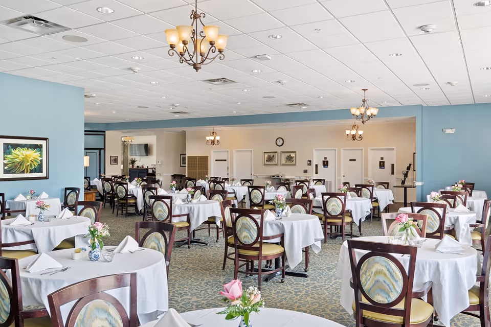 A spacious dining room with multiple round tables covered with white tablecloths, each set with napkins, cutlery, and small floral centerpieces. The room has light blue walls, patterned carpet, and several chandeliers hanging from a white ceiling. There are framed pictures on the walls and doors in the background.