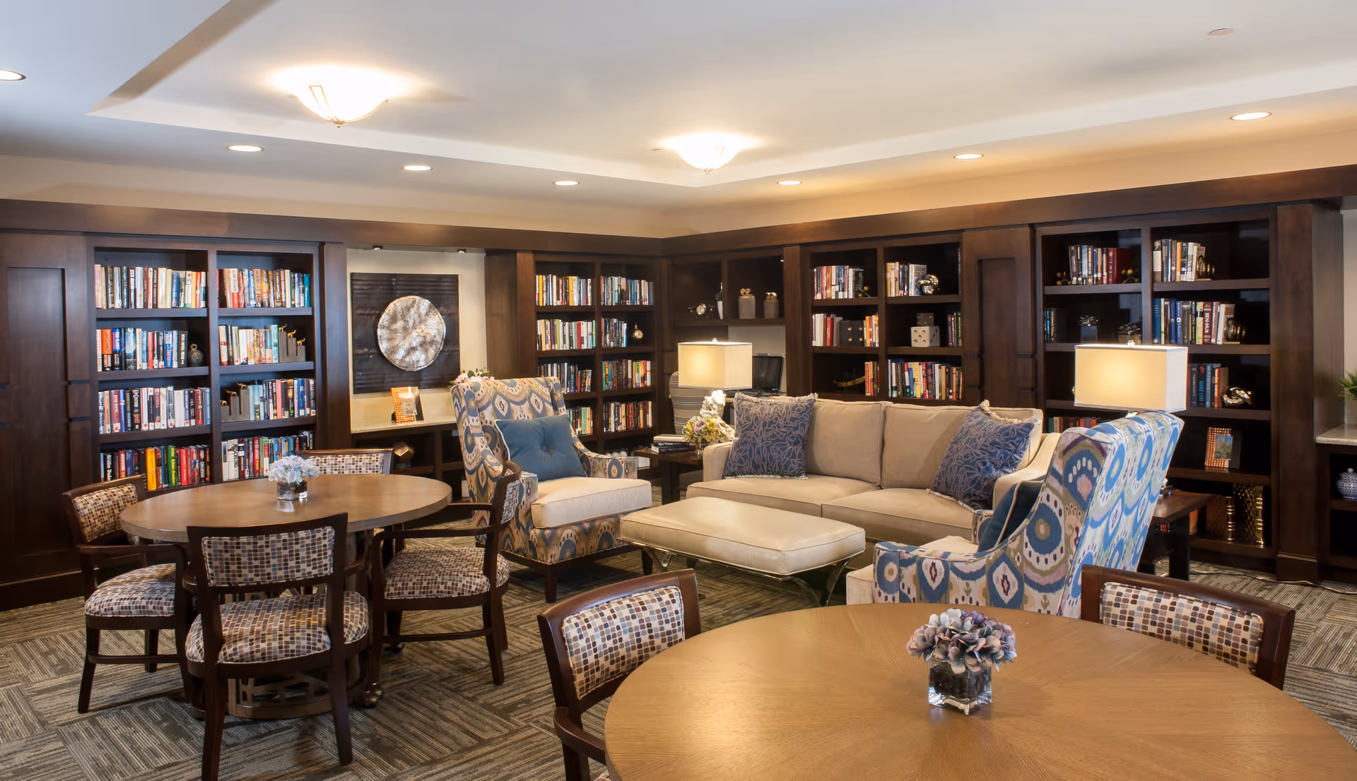Well-lit communal living room and library area with bookshelves, a sofa, patterned armchairs, round tables, and lamps.