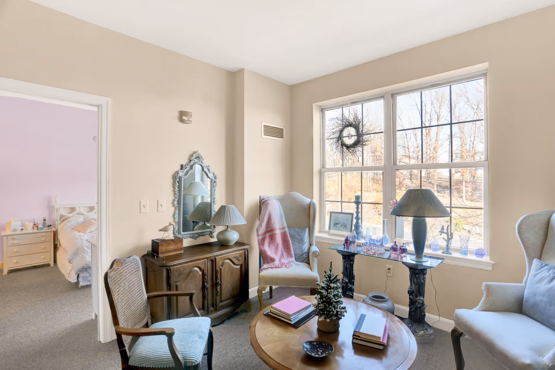 Sunlit sitting room with armchairs, a coffee table and sideboard beside a large window decorated with a wreath, with a bedroom visible through an open doorway.