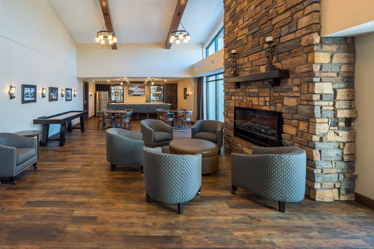 Spacious senior living lounge with curved armchairs around a central ottoman, a stone fireplace on the right, and a dining/bar area in the background.