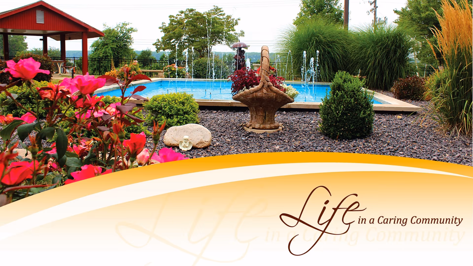 A landscaped outdoor area featuring a small fountain with water jets, surrounded by bushes, flowers, and decorative rocks. In the background, there is a red gazebo with seating and a view of distant trees and hills under a cloudy sky.