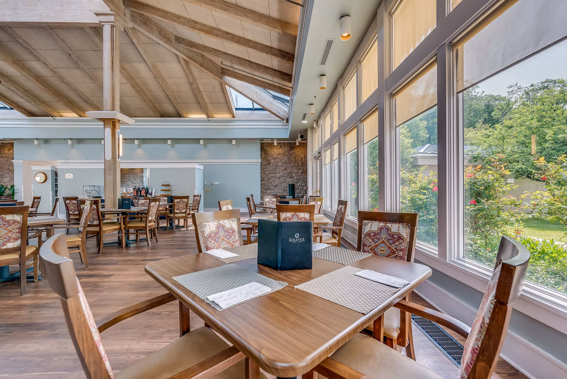 A bright dining room with wooden tables and chairs featuring patterned cushions. Large windows on one side let in natural light and offer a view of a green garden outside. The ceiling has exposed beams and skylights, and the floor is wooden. A menu holder with the word 'Solstice' is placed on the table in the foreground.