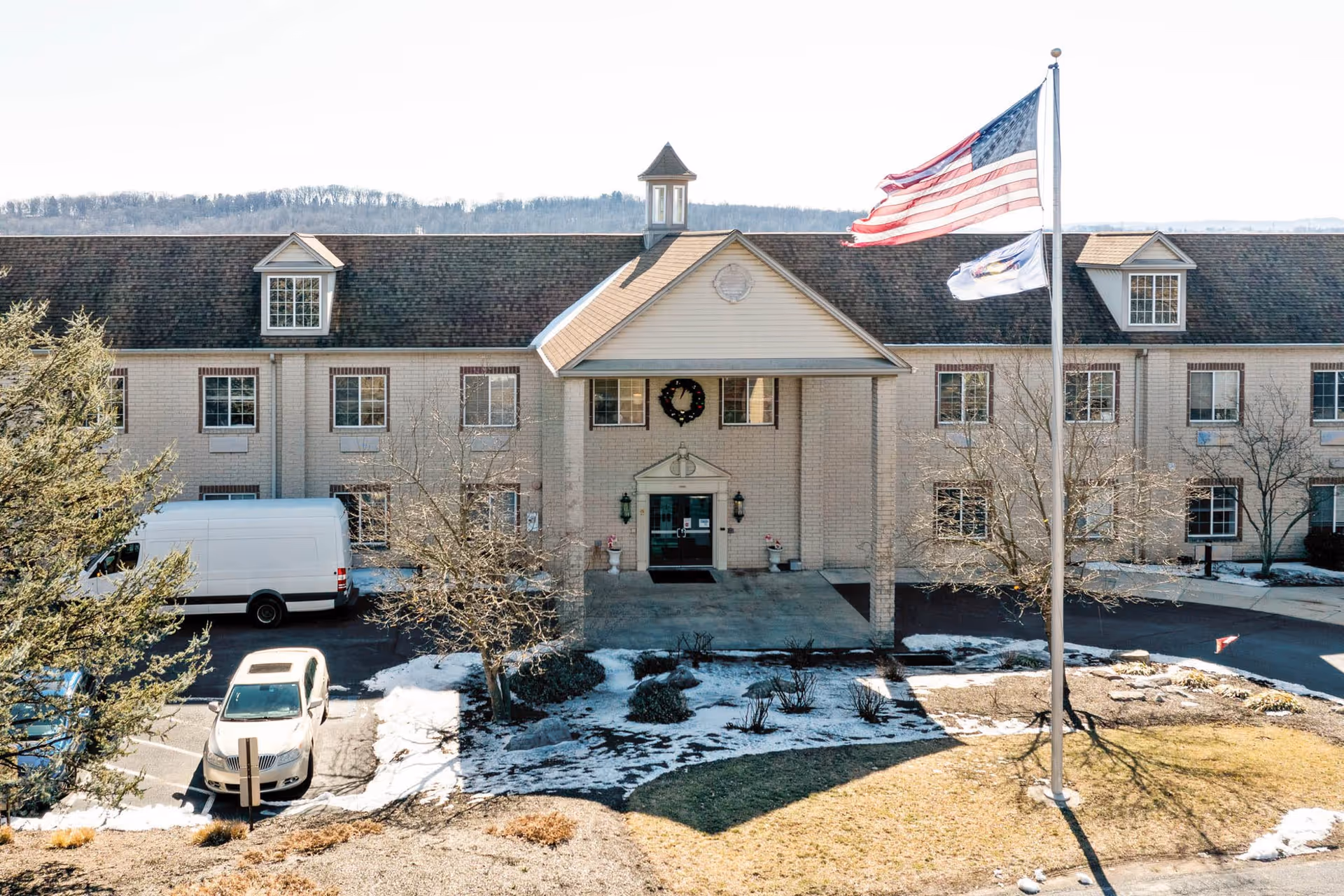 Front exterior view of Greenfield Senior Living at Graysonview building with a covered entrance, American flag and another flag on flagpoles, parked vehicles, some snow on the ground, and trees without leaves.