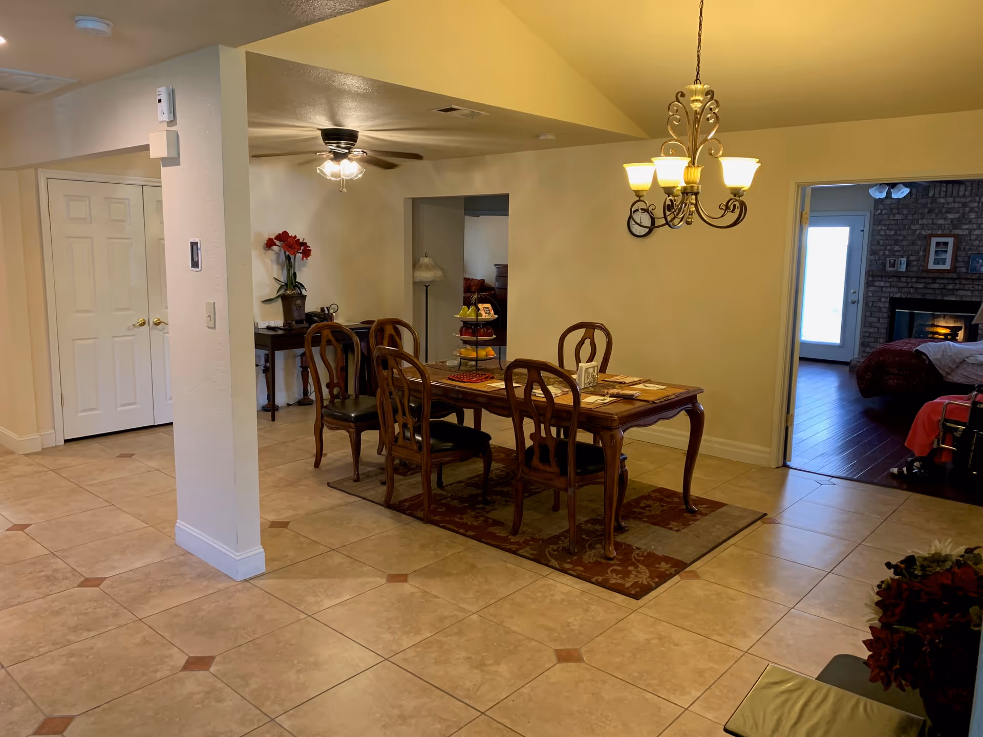Open dining area with a wooden table and six chairs under a chandelier on a tiled floor, adjacent to a living room and hallway.