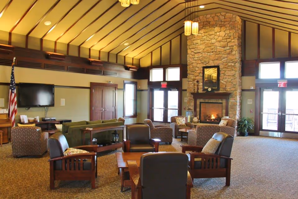 A spacious living room area with a high vaulted ceiling featuring wooden beams. The room includes a stone fireplace with a mirror above it, surrounded by several cushioned armchairs and sofas arranged around a wooden coffee table. There is a flat-screen TV mounted on the wall, an American flag in the corner, and large windows and glass doors letting in natural light.