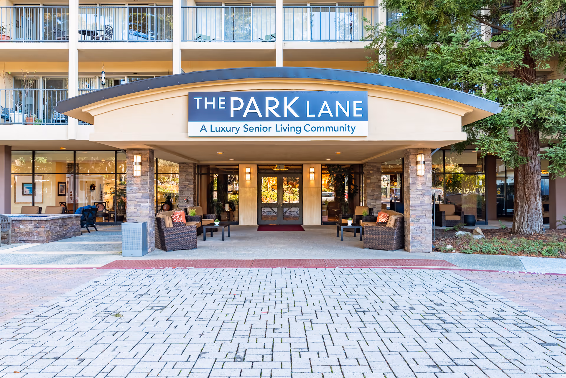 Front entrance of The Park Lane, a luxury senior living community, featuring a covered entryway with stone pillars, outdoor seating with wicker chairs and tables, and a paved driveway leading up to the doors. The building has balconies with railings and large windows.