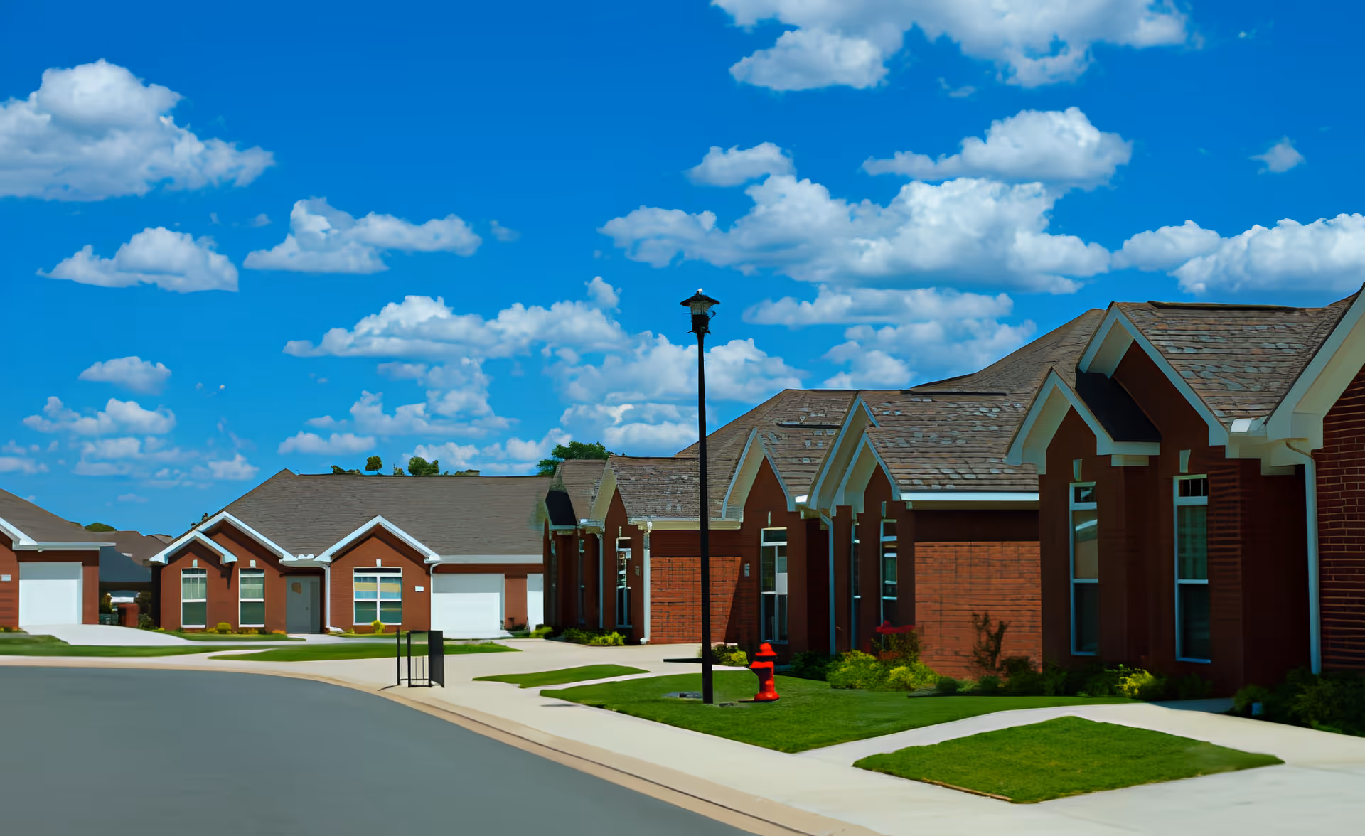 A row of single-story brick residential buildings with peaked roofs along a curved street under a bright blue sky with scattered white clouds. The buildings have large windows and attached garages. There is a street lamp and a red fire hydrant on the grassy area near the sidewalk.