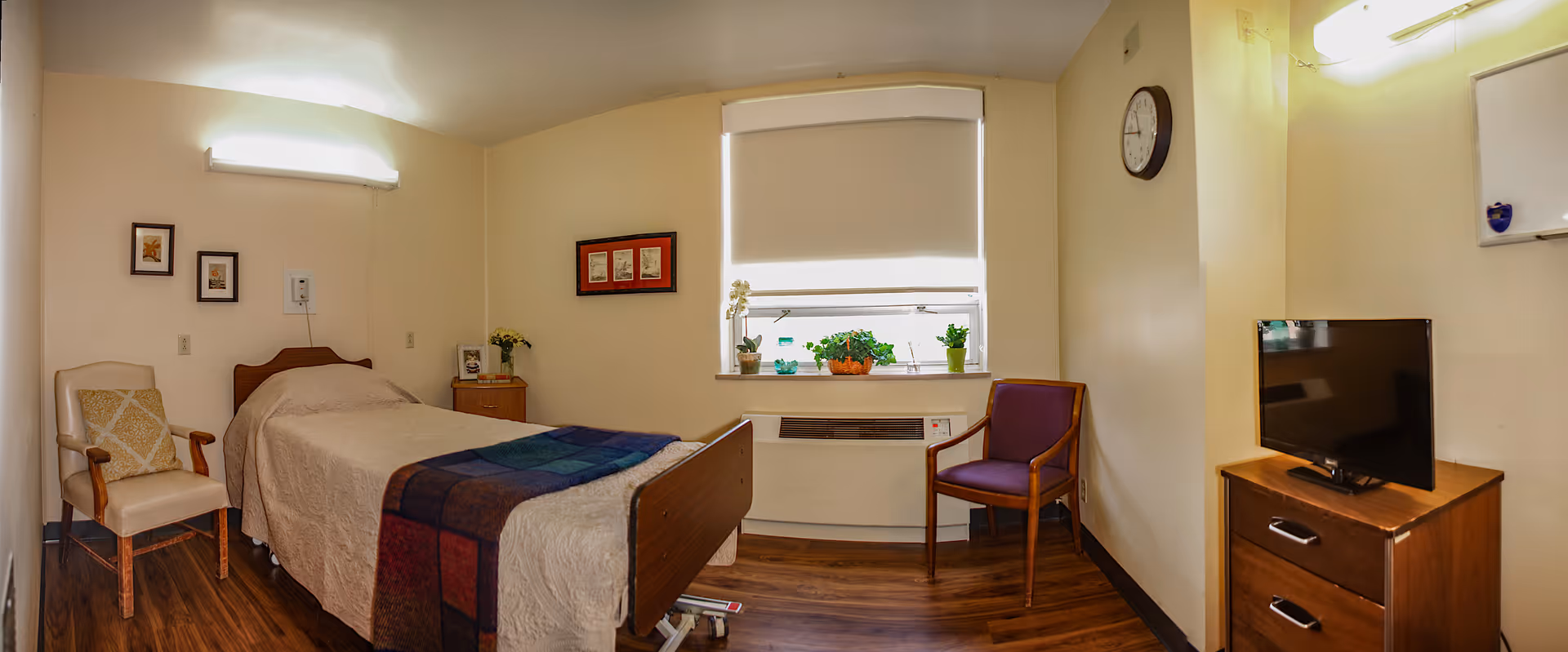A small, cozy bedroom in a rehabilitation facility featuring a single bed with a beige bedspread and a colorful blanket. There are two chairs, one with a cushion and one without, a wooden dresser with a flat-screen TV on top, a window with a white roller shade, and several potted plants on the windowsill. The walls are light-colored with framed pictures and a clock. The floor is wooden.