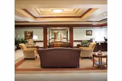 A well-lit communal lounge with sofas, armchairs, side tables, lamps, artwork, and a coffered ceiling.
